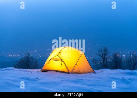 Ein gelbes Zelt, das von innen beleuchtet wird, inmitten des bezaubernden Schimmels der Lichter der Stadt. Eine Darstellung von Reisenden, die in den Winterbergen campen und das Wesen von Reisen und Entdeckungen verkörpern Stockfoto