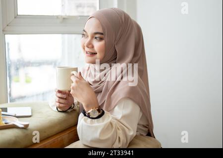 Ein Porträt einer attraktiven jungen asiatischen Frau, die einen Hijab trägt, genießt Kaffee, während sie sich allein in einem Café entspannt. Lifestyle-Konzept Stockfoto