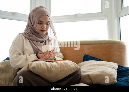 Eine attraktive und fokussierte asiatisch-muslimische Frau genießt ihren Kaffee, während sie im Wohnzimmer ein Buch liest. Wohnkultur und Freizeitkonzepte Stockfoto