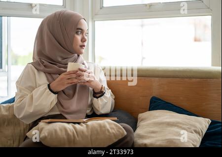 Eine nachdenkliche und charmante junge asiatisch-muslimische Frau, die einen Hijab trägt, eine Kaffeetasse hält, aus dem Fenster blickt und nachdenklich etwas denkt, was was was ist Stockfoto