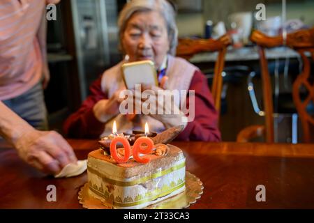 Ältere Frau, die Smartphone-Fotos von einem Kuchen macht, der ihren 90. Geburtstag feiert. Stockfoto