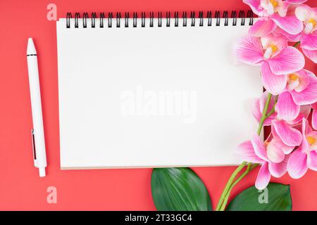 Leeres Notizbuch und Stift mit rosafarbener Orchidee auf rotem Hintergrund. Stockfoto