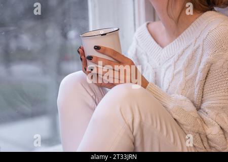 Nahaufnahme weiblicher Hände mit schwarzer Maniküre, die weiße, stilvolle Tasse mit heißem Getränk hält. Winterkakao, Kaffee von Mädchen, das am Fenster sitzt, in weißem strick Stockfoto