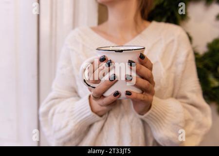 Nahaufnahme weiblicher Hände mit schwarzer Maniküre, die weiße, stilvolle Tasse mit heißem Getränk hält. Winterkakao, Kaffee von Mädchen, das am Fenster sitzt, in weißem strick Stockfoto