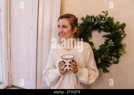 Nahaufnahme von Porträts einer jungen, stilvollen Frau mit weißem Becher in den Händen neben grünem Weihnachtskranz, mit eigenen Händen gemacht. Winterliche festliche Atmosphäre. Florist Stockfoto