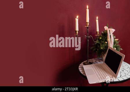 Hintergrund der roten weinroten Wand, weißer Laptop mit Mini-Weihnachtsbaum auf dem Tisch. Working Holiday Space. Platz für Text. Stilvolle, moderne Einrichtung. Soci Stockfoto