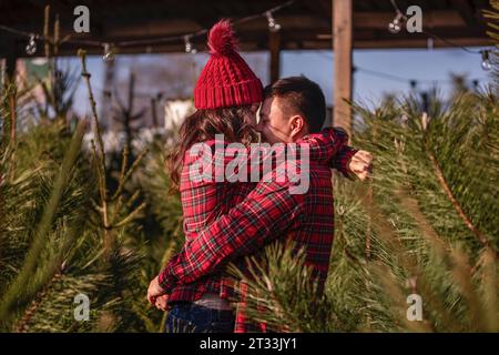 Wunderschönes Paar in karierten roten Hemden, Strickmützen umarmen sich von Angesicht zu Angesicht zwischen grünem Weihnachtsbaummarkt. Junger Mann lacht Stockfoto
