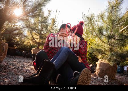 Rot-weißer Zuckerrohr wird in den ausgestreckten Händen des liebenden Paares in karierten Hemden gehalten, Strickmützen in der Nähe des grünen Weihnachtsmarktes. Glücklicher Mann Stockfoto