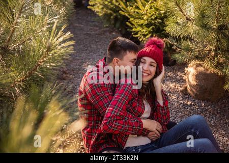 Nahaufnahmen von jungen Paaren, die sich im Wald entspannen und rote karierte Hemden tragen, die auf dem Straßenmarkt zwischen Weihnachtsbaumkeimlingen sitzen. Mann Embra Stockfoto