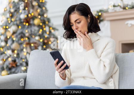Verärgerte Frau, die allein zu Hause auf dem Sofa in der Nähe des Weihnachtsbaumes sitzt, Frau, die Silvester und Weihnachten feiert, Telefon hält, Online-Benachrichtigung mit schlechten Nachrichten, unglückliches Lesen erhielt. Stockfoto