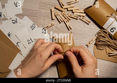 Eine nicht wiedererkennbare junge Frau klebt die Nummer auf die Handwerkstasche, wird mit einer Wäscheklammer befestigt. Weiblich macht Kraftpapier für hausgemachte Adventskalender mit eigenen Händen Schritt für Schritt selbst gemacht. Vorbereitung auf das weihnachtskonzept. Saisonale Aktivitäten für Kinder, Familienurlaub im Winter. Umweltfreundlich Geschenke. Öffnen Sie das Paket jeden Tag Stockfoto