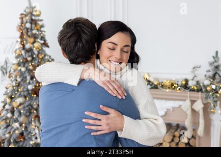 Familienpaar feiert gemeinsam Silvester an Weihnachten, steht neben dem Weihnachtsbaum im Wohnzimmer, lächelt fröhlich und umarmt, Mann und Frau in l Stockfoto