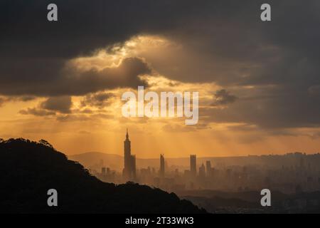 In der Abenddämmerung bricht die Sonne durch die Wolken und scheint auf Taipeh City. Silhouetten von Stadtgebäuden. Orangefarbener Himmel und dunkle Wolken. Stockfoto