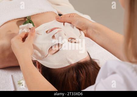 Kosmetikerin glättet Bettmaske auf Frau Gesicht für Rehydrate Gesicht Haut, Anti Falten kosmetische Prozedur im Beauty Spa Salon. Kosmetologe, der moi anwendet Stockfoto