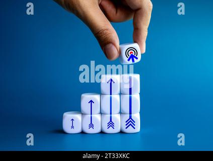 Das Zielsymbol wird von der Hand des Geschäftsmannes gehalten und auf den weißen Würfel-Block gelegt, während der Wachstumsdiagramm mit aufsteigenden Pfeilsymbolen auf blauem Hintergrund verläuft. Ziel A Stockfoto