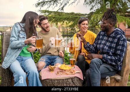 Vier Erwachsene sitzen draußen an einem Baumstamm-Tisch und teilen sich Lachen und Bier. Snacks werden verteilt, während sie die Gesellschaft des anderen in einem Gartenset genießen Stockfoto