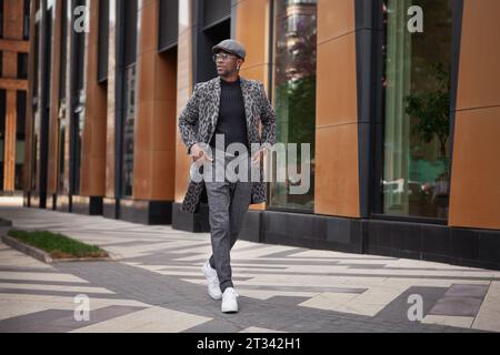 Modemann mit modischer Kleidung grauer Mantel mit Tierdruck, Rollkragen, Mütze, Hose und weißen Sneakers. Im Freien. Männliches stilvolles Modell, das sich auf dem Laufenden befindet Stockfoto