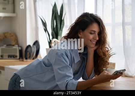 Glückliche, angenehme junge Frau, die mit dem Smartphone in der Küche steht und online nach Kochrezepten sucht Stockfoto