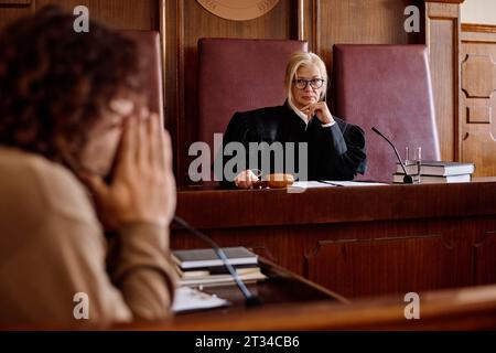 Ernster blonder, unparteiischer Richter in Brille und Mantel, der jungen männlichen Verdächtigen ansieht, während er am Schreibtisch sitzt und seinen Aussagen zuhört Stockfoto