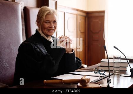 Reifer, erfolgreicher blonder Richter im schwarzen Mantel, der mit einem Lächeln in die Kamera blickt, während er vor den Mikrofonen im Gerichtssaal am Schreibtisch sitzt Stockfoto