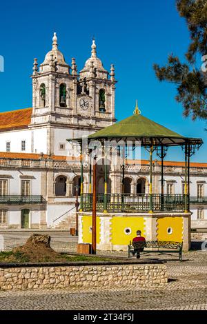 Nazaré, Portugal - Februar 2023: Der Altstädter Platz von Nazaré mit einem gelben Bühnenpult und einem Mann, der auf einer Bank vor der Kirche Nossa Senhora da Nazaré sitzt. Stockfoto