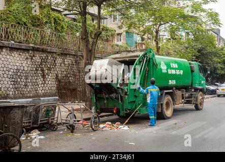 Ein Müllmann in blauer Uniform betreibt einen automatisierten Müllwagen neben traditionellen Handkarren im Zentrum von Hanoi, Vietnam. Stockfoto