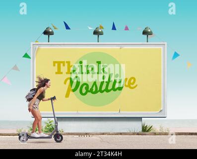 Glückliche Frau, die einen E-Scooter fährt und an den Strand geht, motivierendes Poster im Hintergrund: Denken Sie positiv Stockfoto