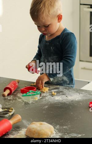 Kleiner kleiner Junge, der zu Hause Weihnachtskekse backt. Roter Pin- und Keksschneider auf dunkelgrauem Küchentisch. Stockfoto