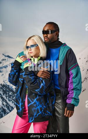 Wunderschönes Paar aus verschiedenen Rassen in lebhaften, auffälligen Outfits und stilvollen Sonnenbrillen, die sich im Winter warm umschmeicheln Stockfoto