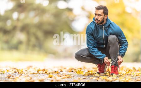 Ein junger Athlet in warmer Sportbekleidung bindet seine Schnürsenkel, während er in einem Herbstpark läuft. Stockfoto