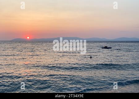 Sonnenuntergang auf dem ägäischen Mittelmeer mit Pinienbäumen, die sich hinter den Bergen am Horizont sonnen. Menschen schwimmen und auf schwimmenden Pontonbahnen Stockfoto