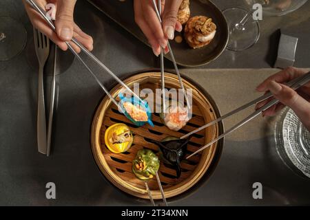 Die Hände verschiedener Personen verwenden Essstäbchen, um eine Vielzahl traditioneller gedämpfter Dim Sum aus einem Bambus-Dampfgarer zu pflücken, die alle elegant auf einer dunklen Tafel serviert werden Stockfoto