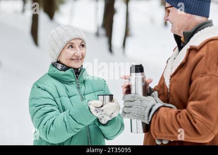 Lächelnde Seniorin bittet den Ehemann, ihren heißen Tee aus der Thermoskanne zu gießen Stockfoto