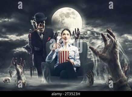 Eine verängstigte Frau, die Horrorfilme ansieht und Popcorn isst, sie ist von gruseligen Zombiehänden umgeben und ein Skelettmonster steht hinter ihr Stockfoto