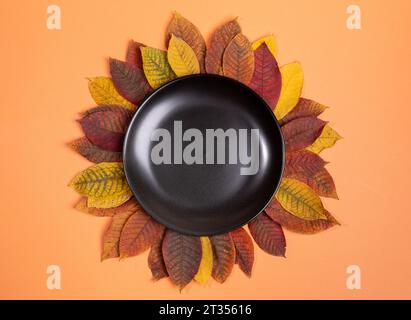 Schwarze runde Platte mit bunten Herbstblättern auf orangefarbenem Hintergrund. Herbstmotiv. Herbst, Halloween, Thanksgiving Tag Layout. Draufsicht. Kopieren Stockfoto