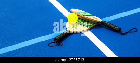 West Islip, New York, USA - 17. Juli 2023: Zwei Pickleball-Paddel mit einem blühenden Whistle Ball auf zwei Linien auf einem blauen Pickleball-Tennisplatz. Stockfoto