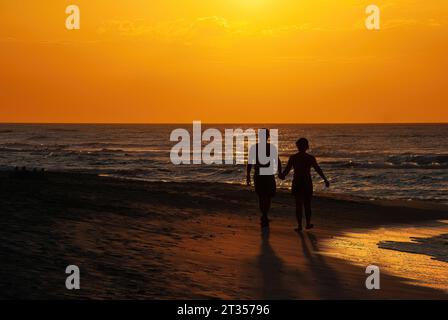 Silhouette eines Paares, das Hände hält, während man bei Sonnenuntergang am Strand spaziert, sehr romantisch Stockfoto