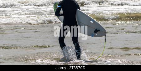Rückansicht eines Mannes in einem schwarzen Neoprenanzug, der mit seinem Surfbrett ins Meer geht. Stockfoto