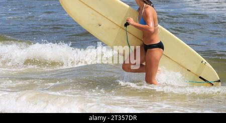 Seitenansicht eines jungen Surfermädchens, das einen Bikini trägt und ihr Surfbrett aus der Nähe aus dem Meer trägt. Stockfoto