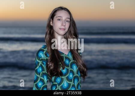 Eine 20-jährige Kaukasierin steht bei Sonnenuntergang am Strand und trägt ein lebendiges und farbenfrohes Hemdkleid, während die Sonne ein warmes Licht über den Strand wirft Stockfoto