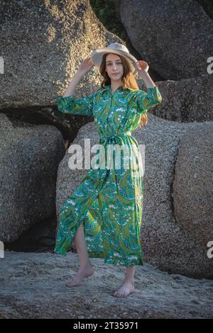 Eine 20-jährige Kaukasierin strahlt am Strand natürliche Schönheit aus, die mit einem grünen Hemdkleid und einem Hut vor einem Hintergrund von Felsbrocken verziert ist Stockfoto