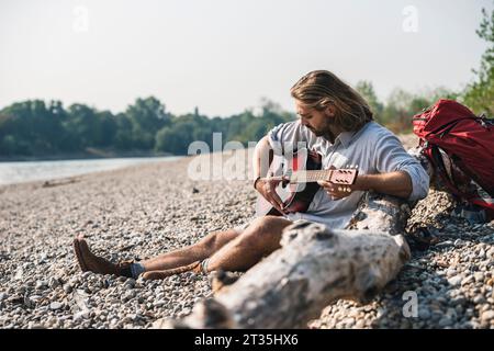Junger Mann am Flußufer Gitarre spielen Sitzen Stockfoto