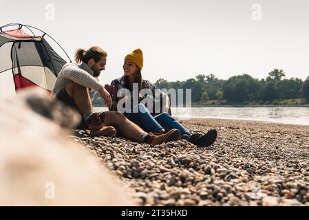 Glückliches junges Paar mit Gitarre sitzt an einem Zelt am Flußufer Stockfoto