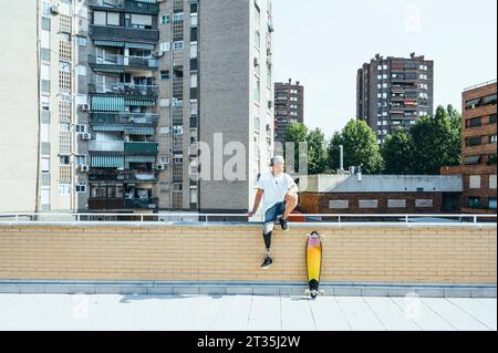 Junger Mann mit Bein Prothese sitzt auf Geländer in der Stadt neben Skateboard Stockfoto