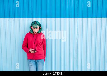 Junge Frau mit blau gefärbtem Haar hört Musik mit Kopfhörern, während sie auf das Smartphone blickt Stockfoto