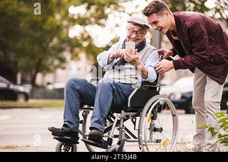 Lachend älterer Mann im Rollstuhl und seines erwachsenen Enkel zusammen am Smartphone Spaß suchen Stockfoto