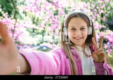 Lächelndes Teenager-Mädchen mit Friedenszeichen und Musik mit Kopfhörern im Park Stockfoto