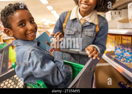 Lächelnder Junge, der im Einkaufswagen mit Mutter im Supermarkt sitzt Stockfoto