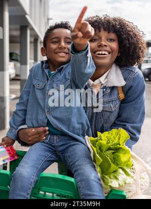 Glückliche Frau mit einem Jungen, der im Einkaufswagen sitzt und Gesten macht Stockfoto