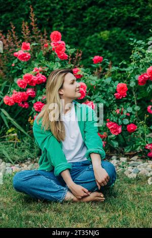 Lächelnde Frau, die vor Rosenpflanzen im Garten sitzt Stockfoto